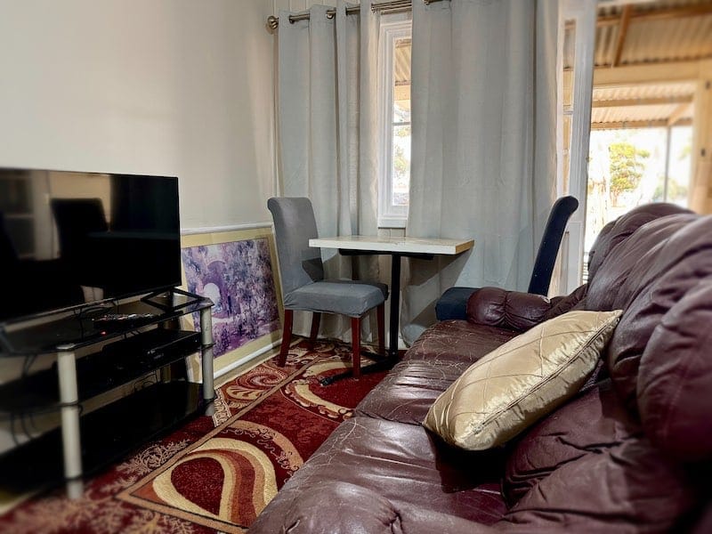 The living area. It has a TV, a comfortable sofa, and a soft red rug on the floor. There is also a small dining table by the window, with two chairs.